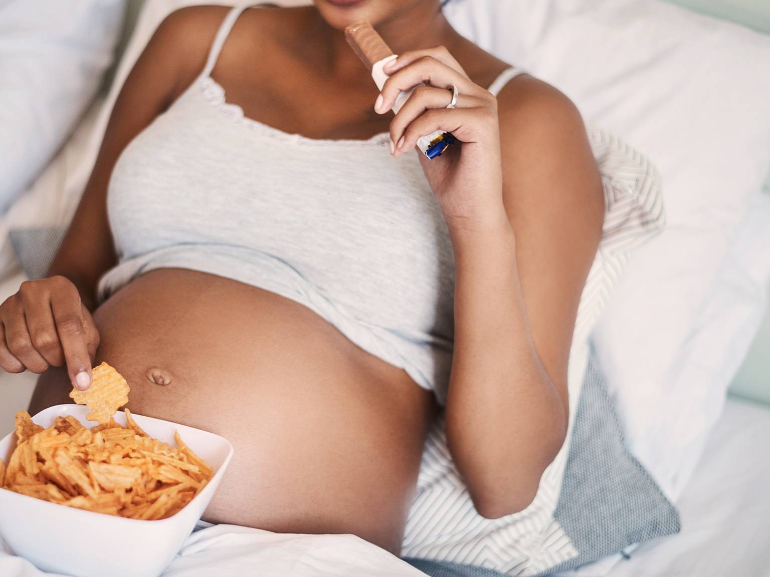 Schwangere liegt im Bett und isst Chips