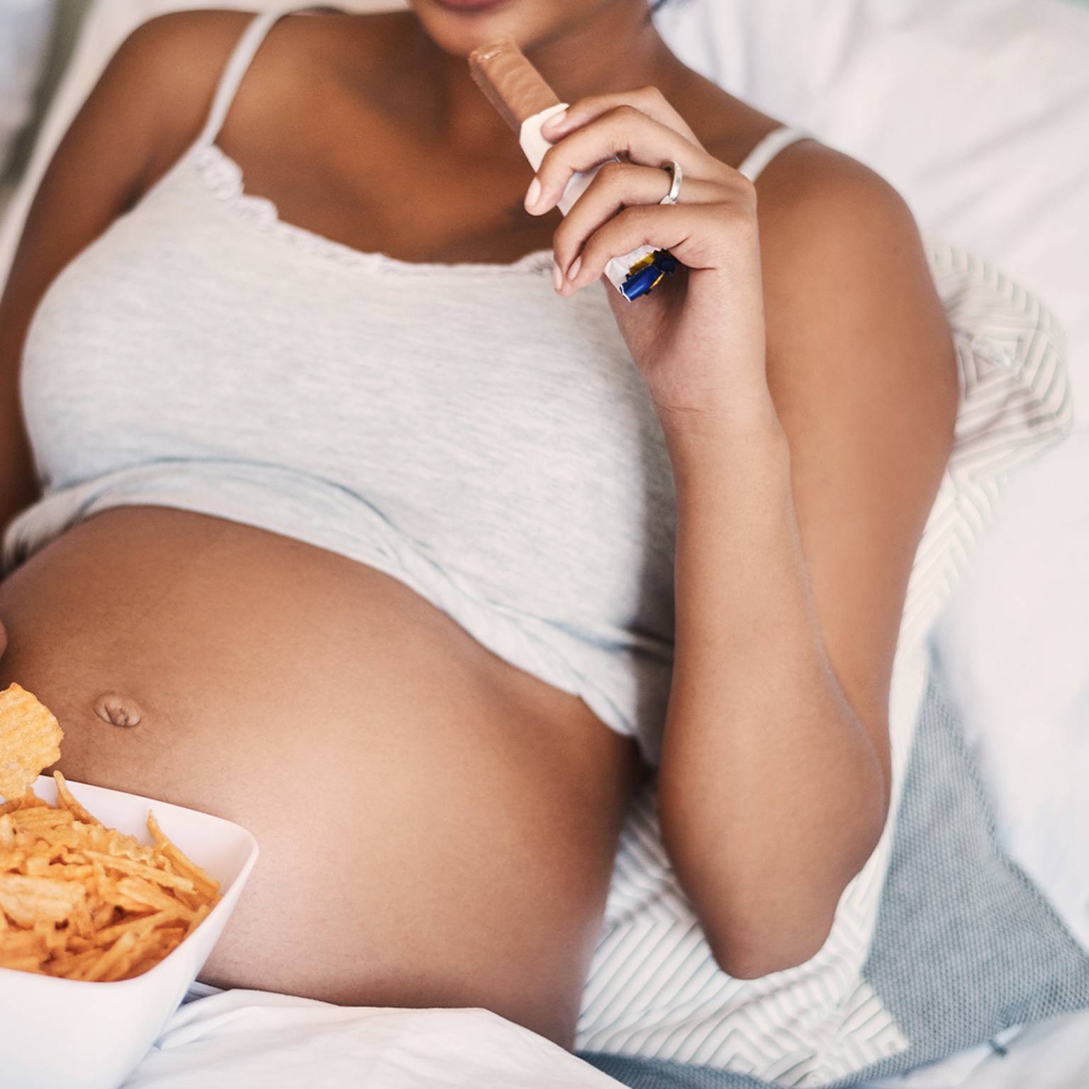 Schwangere liegt im Bett und isst Chips