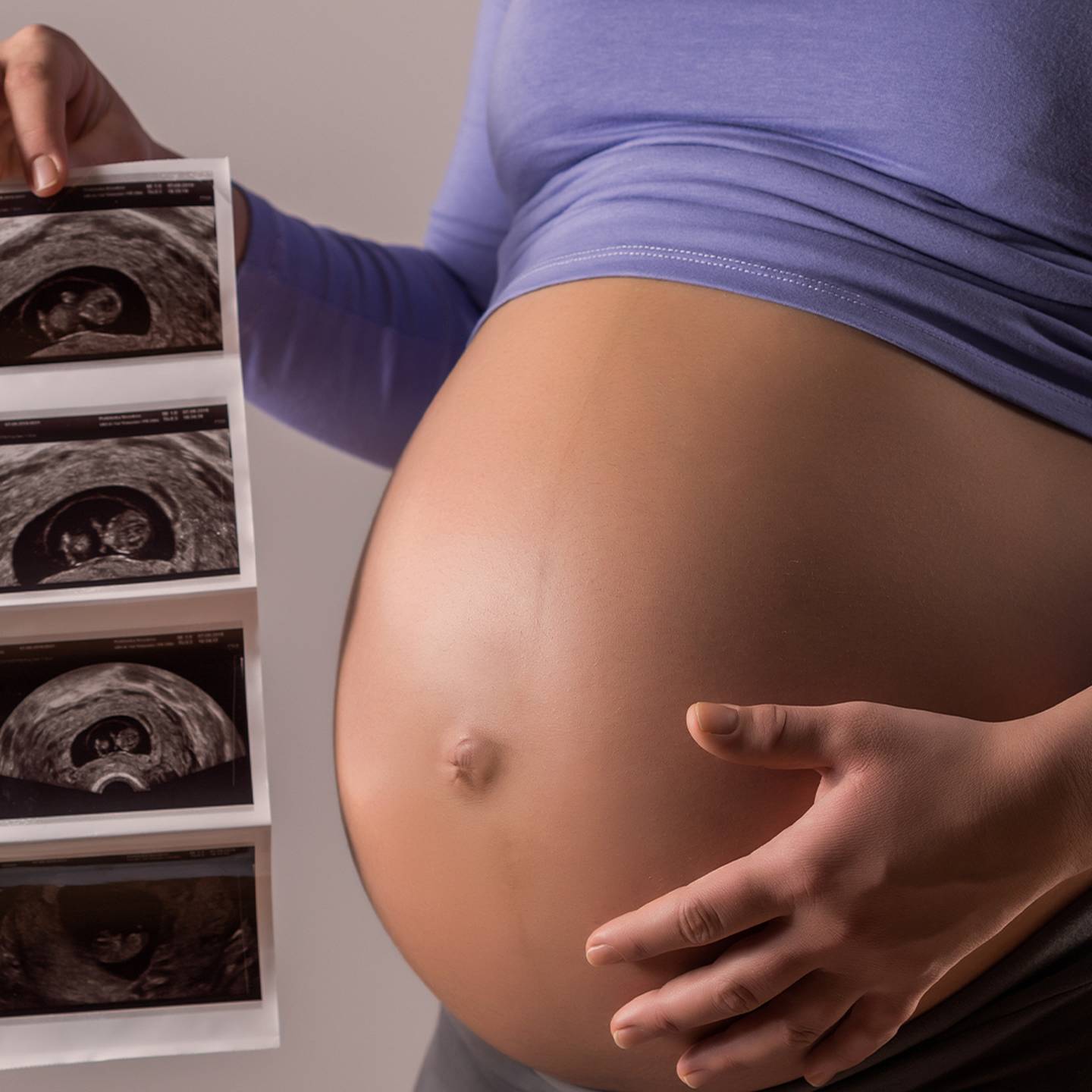 Schwangere Person, die Ultraschallbilder an ihren nackten Bauch hält und ein blaues Oberteil und eine graue Hose trägt.