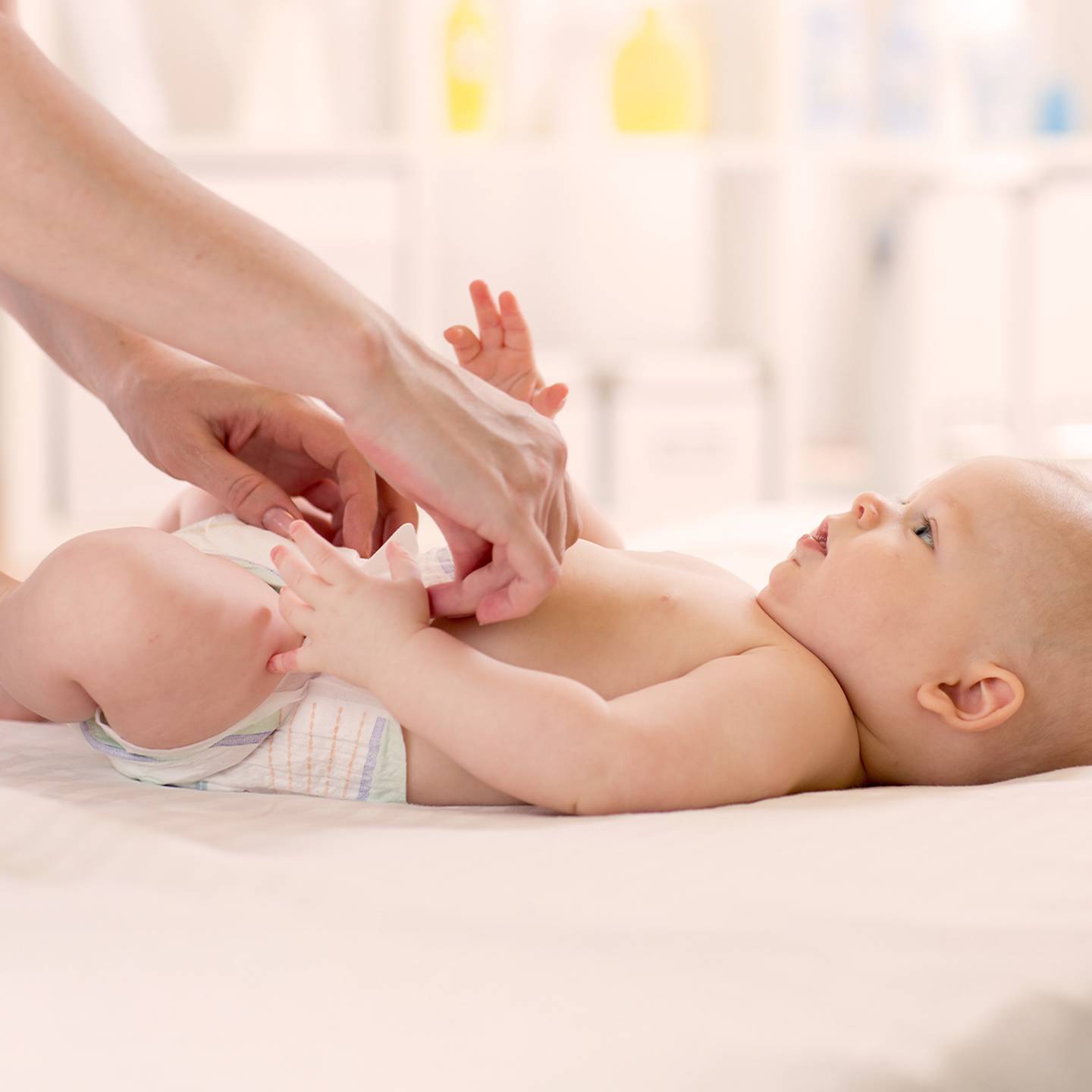 Ein Erwachsener wechselt die Windel eines Babys auf einem weißen Wickeltisch in einem hellen Raum. Das Baby blickt nach oben und hat die Arme leicht erhoben.