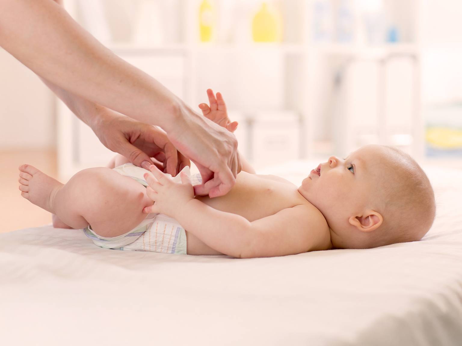 Ein Erwachsener wechselt die Windel eines Babys auf einem weißen Wickeltisch in einem hellen Raum. Das Baby blickt nach oben und hat die Arme leicht erhoben.