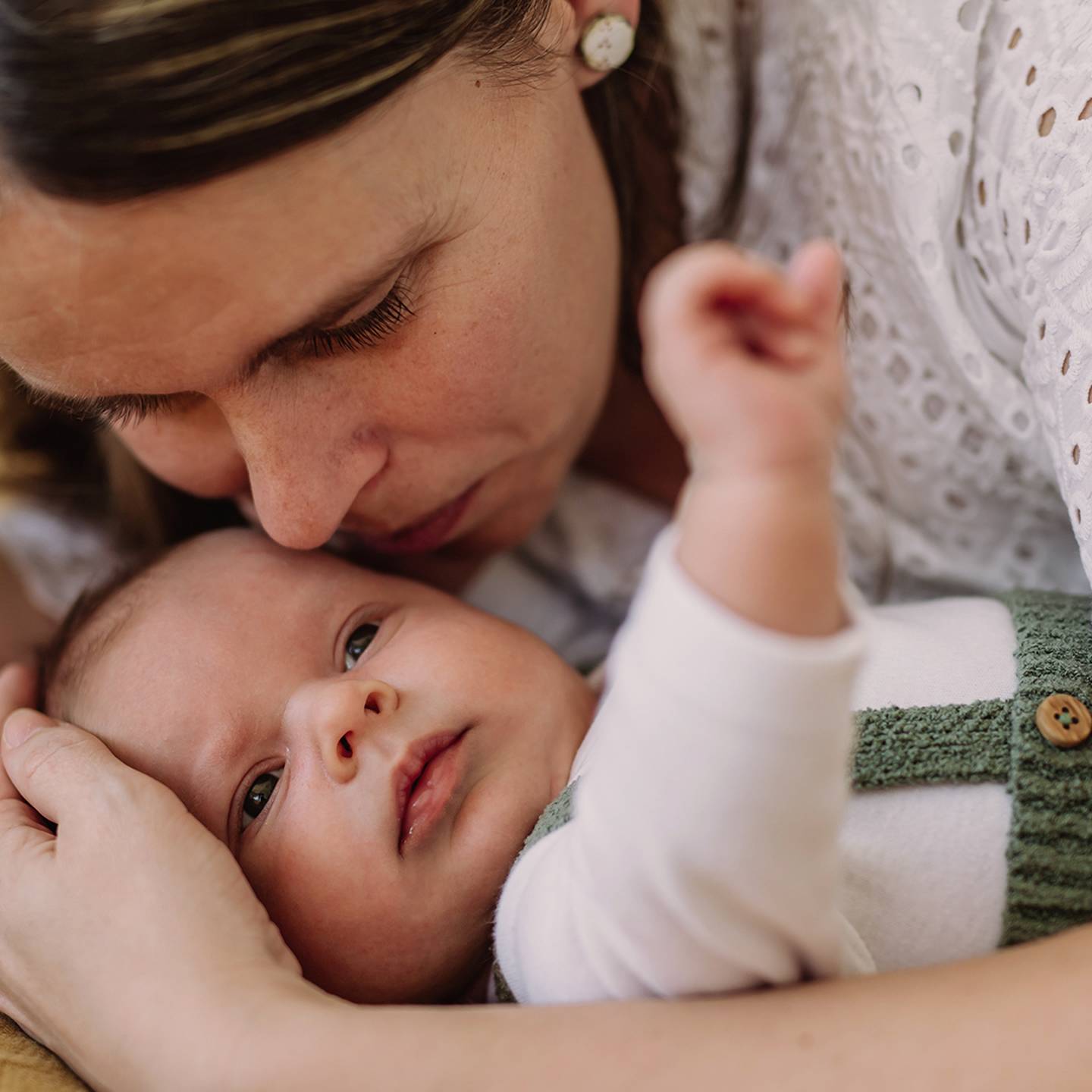 Eine Frau umarmt liebevoll ein Baby. Die beiden liegen eng aneinander gekuschelt auf einer weichen Unterlage. Das Baby blickt nach oben und trägt ein weißes Outfit mit grünen Trägern.