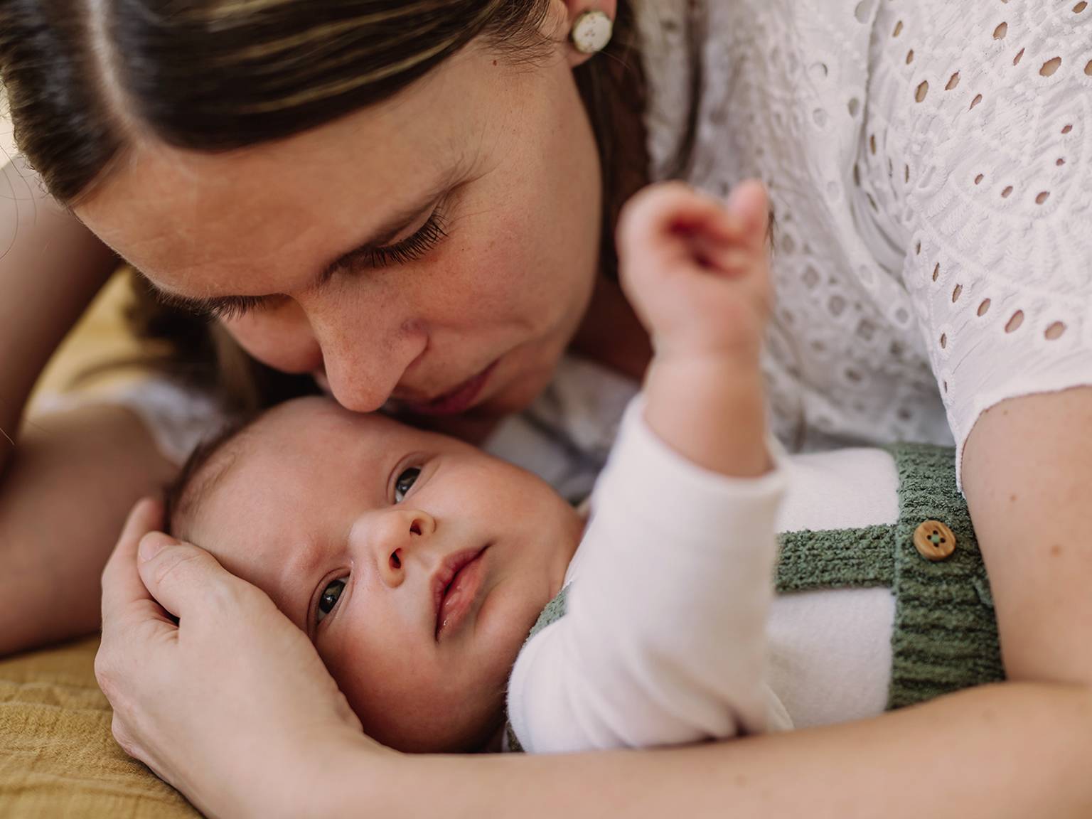 Eine Frau umarmt liebevoll ein Baby. Die beiden liegen eng aneinander gekuschelt auf einer weichen Unterlage. Das Baby blickt nach oben und trägt ein weißes Outfit mit grünen Trägern.