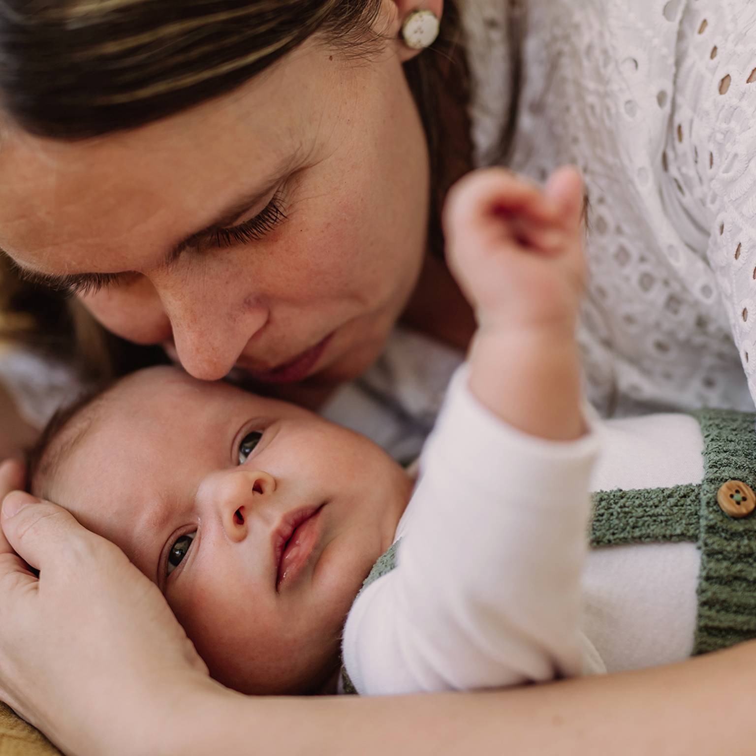 Eine Frau umarmt liebevoll ein Baby. Die beiden liegen eng aneinander gekuschelt auf einer weichen Unterlage. Das Baby blickt nach oben und trägt ein weißes Outfit mit grünen Trägern.