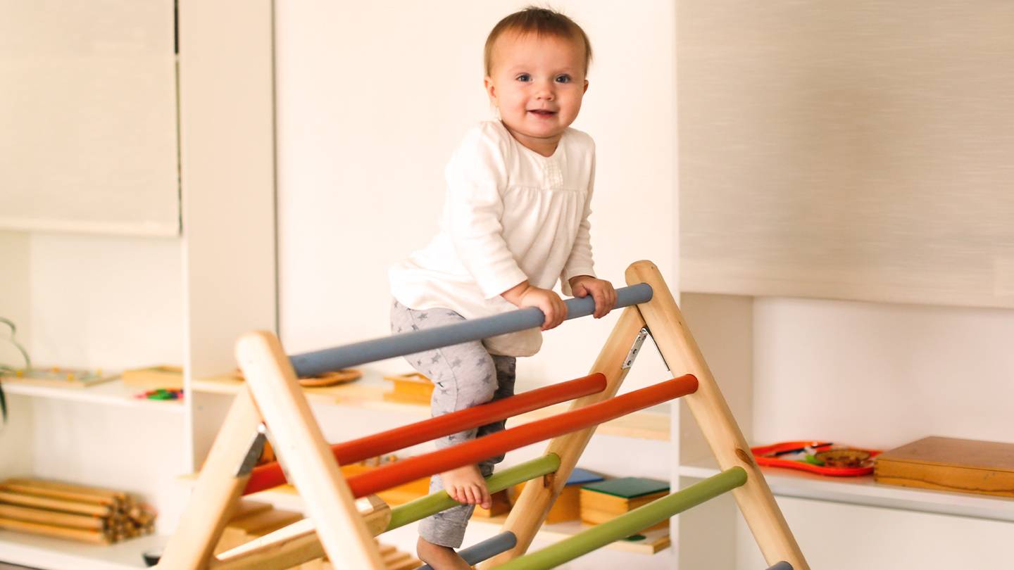Ein lächelndes Kleinkind klettert in einem weißen Hemd und einer grauen Hose auf ein buntes Holzspielgerät im Haus. Im Hintergrund sind Regale mit Spielzeug zu sehen.