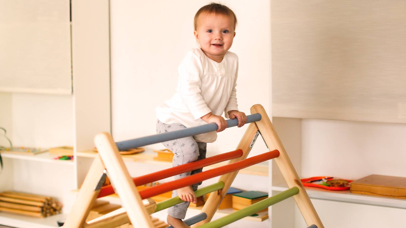Ein lächelndes Kleinkind klettert in einem weißen Hemd und einer grauen Hose auf ein buntes Holzspielgerät im Haus. Im Hintergrund sind Regale mit Spielzeug zu sehen.