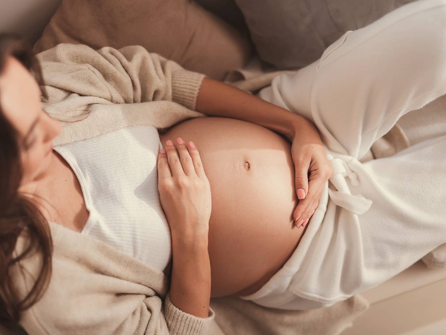 Schwangere Frau in gemütlicher Kleidung, die auf einer Couch liegt und mit beiden Händen sanft ihren Bauch berührt, was Ruhe und Zufriedenheit ausdrückt.