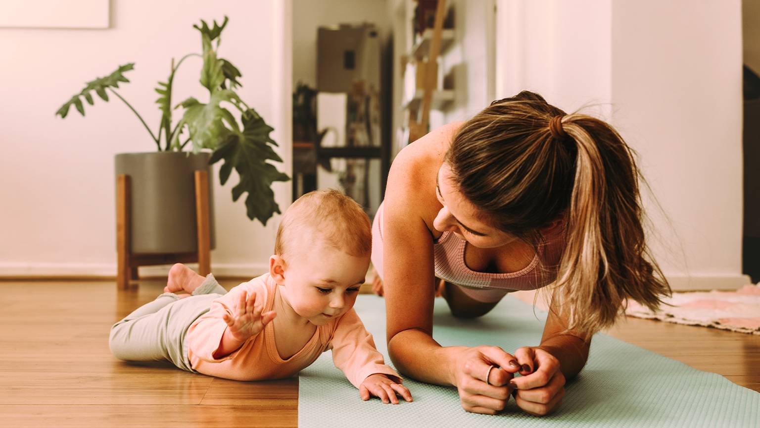 Frau macht eine Plank-Übung auf einer Yogamatte und lächelt ein Baby an, das neben ihr auf dem Boden in einem gemütlichen Zimmer mit einer Topfpflanze liegt.