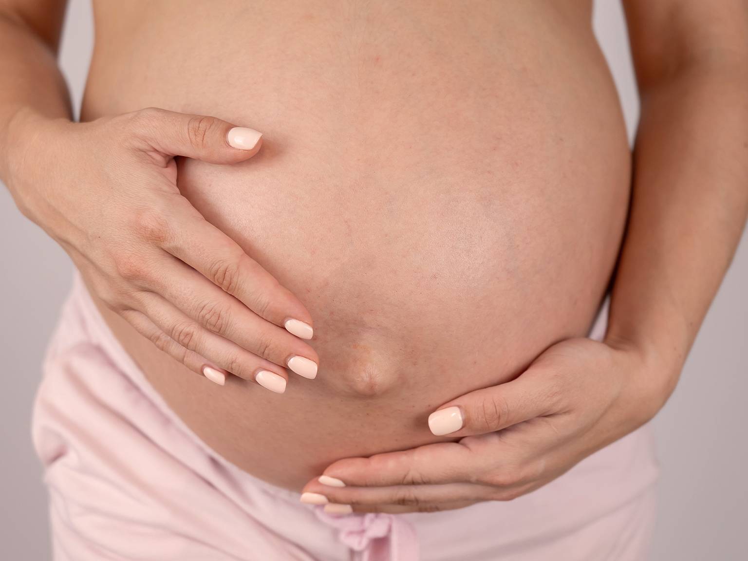 Nahaufnahme einer schwangeren Person, die ihren nackten Bauch mit beiden Händen hält und eine hellrosa Hose trägt.