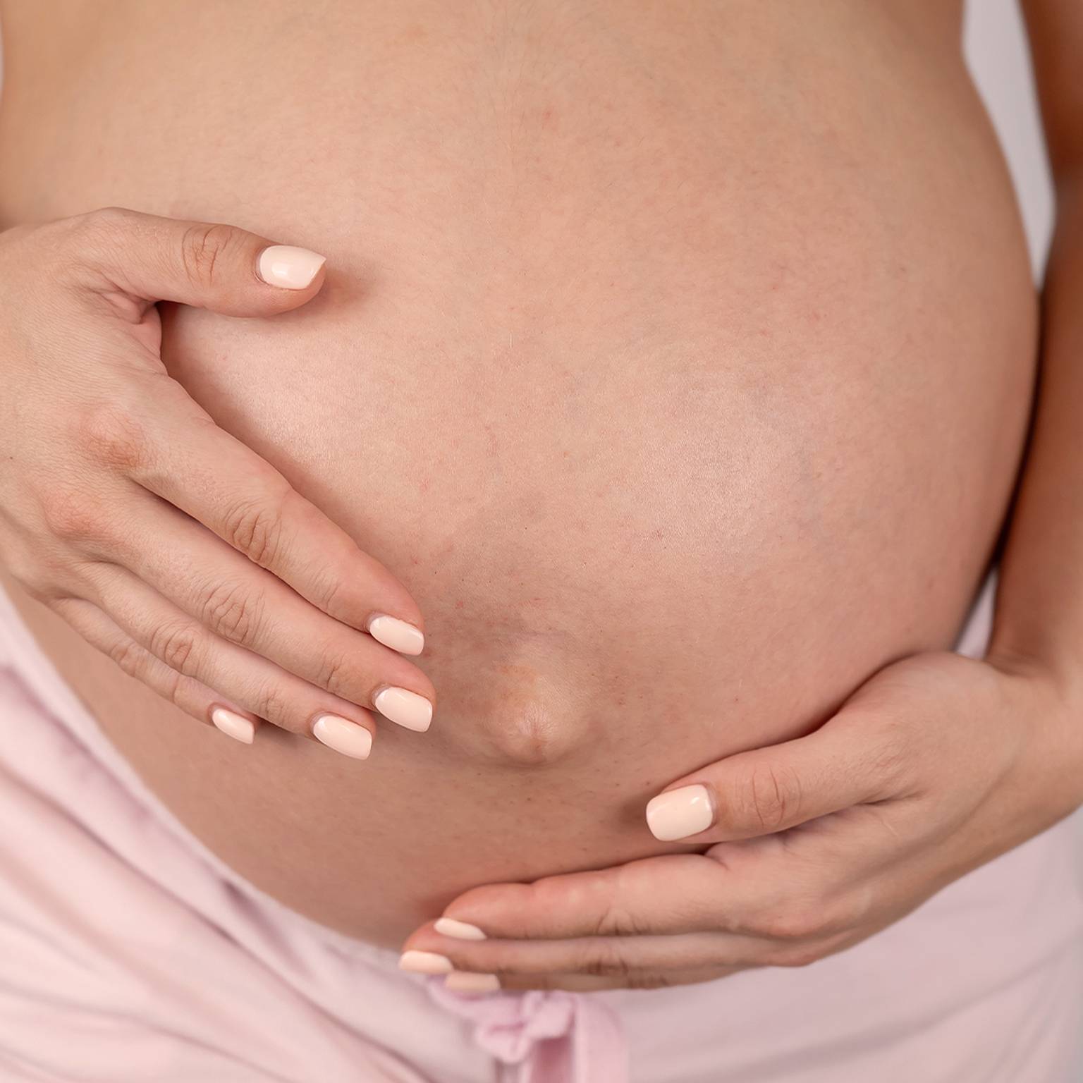 Nahaufnahme einer schwangeren Person, die ihren nackten Bauch mit beiden Händen hält und eine hellrosa Hose trägt.