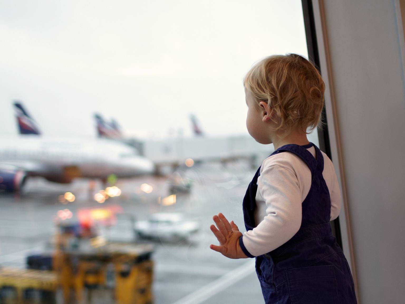 Ein kleines Kind im Overall schaut aus einem Flughafenfenster und beobachtet im Hintergrund Flugzeuge auf dem Rollfeld.