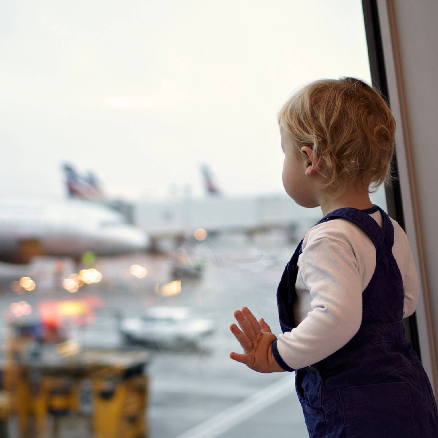 Ein kleines Kind im Overall schaut aus einem Flughafenfenster und beobachtet im Hintergrund Flugzeuge auf dem Rollfeld.