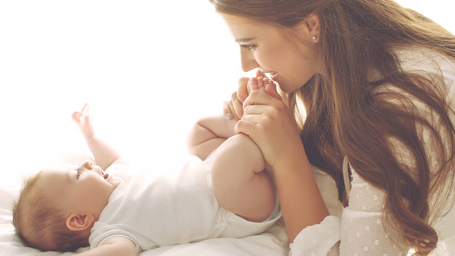 Eine lächelnde Frau spielt sanft mit einem Baby, das auf einem Bett liegt, und beide schauen sich in einem hellen, sanft beleuchteten Raum freudig an.