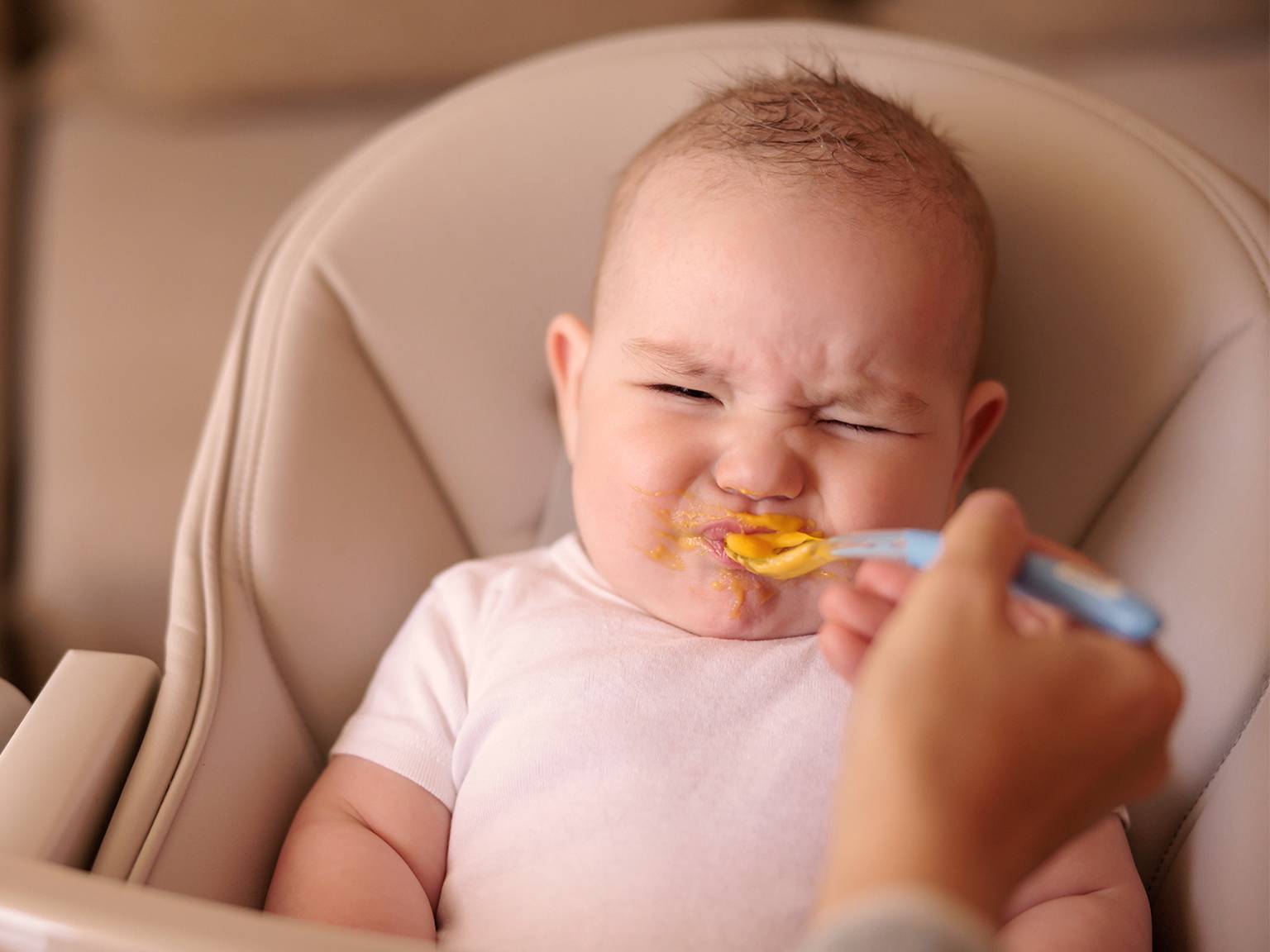 Baby im Hochstuhl, das Grimassen schneidet, während es mit einem Löffel orangefarbener Babynahrung gefüttert wird.