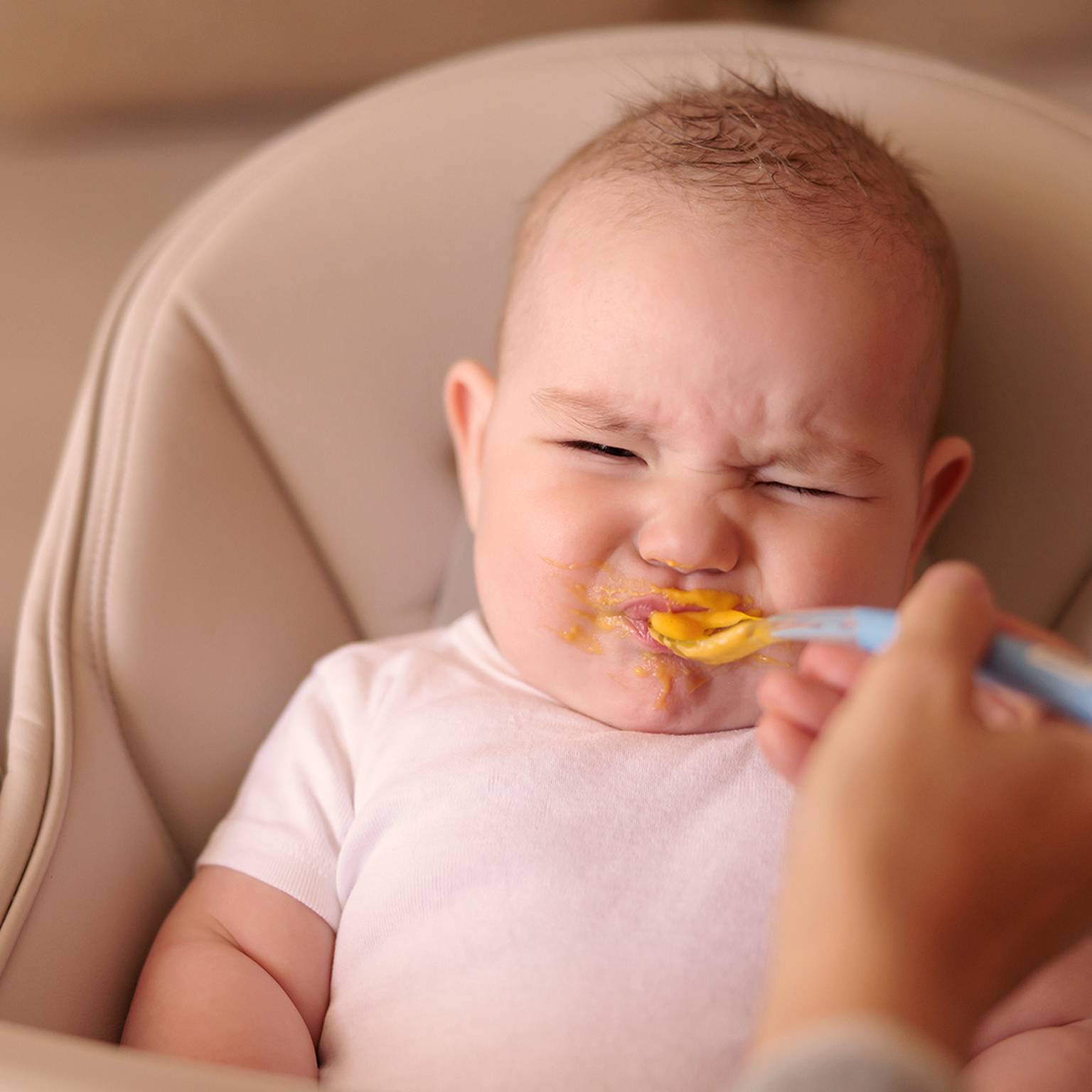 Baby im Hochstuhl, das Grimassen schneidet, während es mit einem Löffel orangefarbener Babynahrung gefüttert wird.