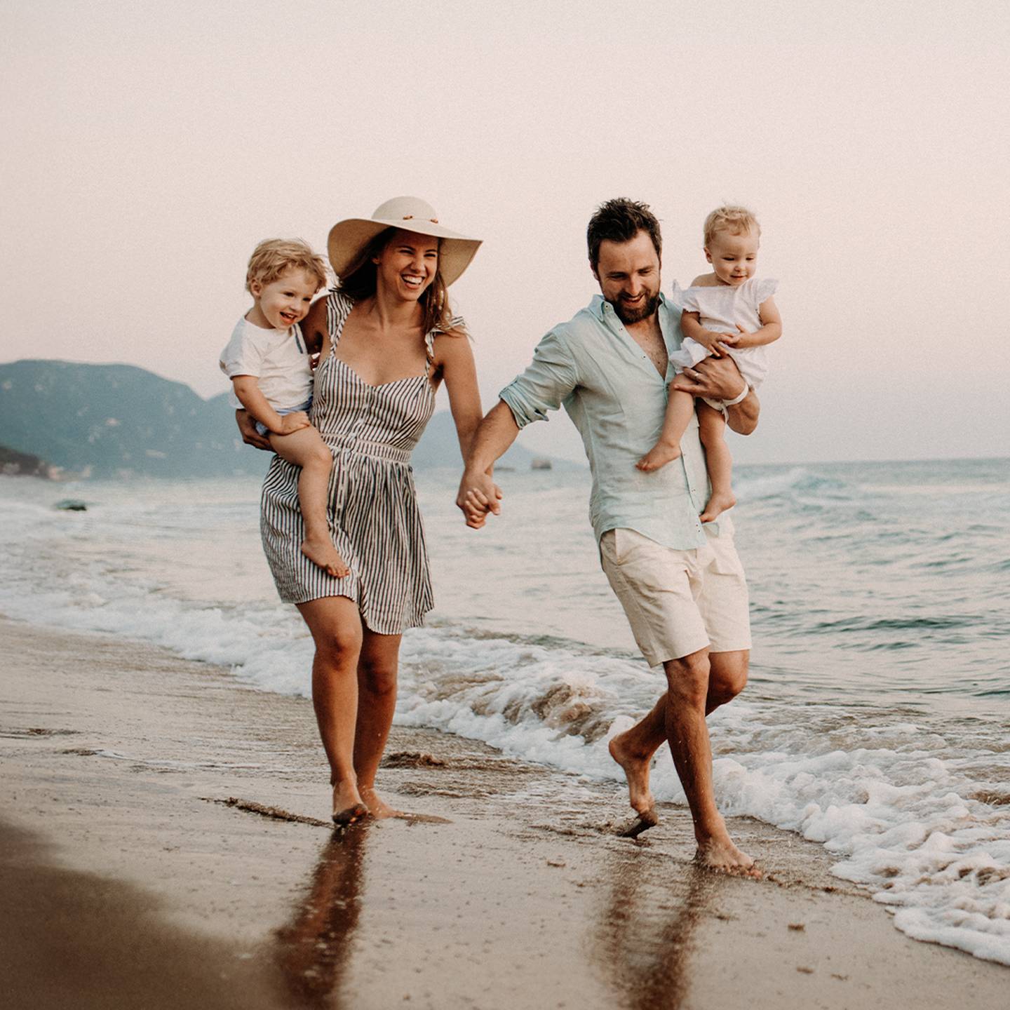Eine vierköpfige Familie läuft barfuß an einem Sandstrand, die Eltern halten ihre Kinder, im Hintergrund sanfte Wellen und Berge bei Sonnenuntergang.