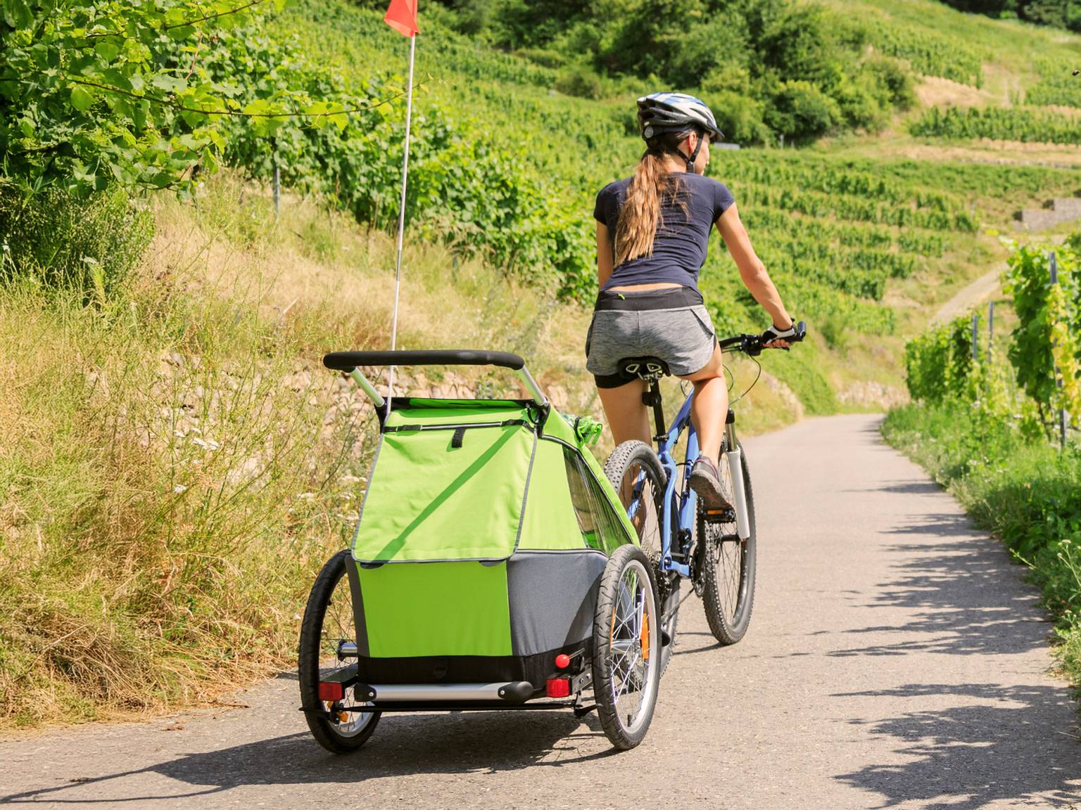 Eine Frau fährt mit dem Fahrrad auf einem Weg durch üppige Weinberge und zieht einen grünen Kinderanhänger hinter sich her. Sie trägt einen Helm und lässige Sportkleidung.