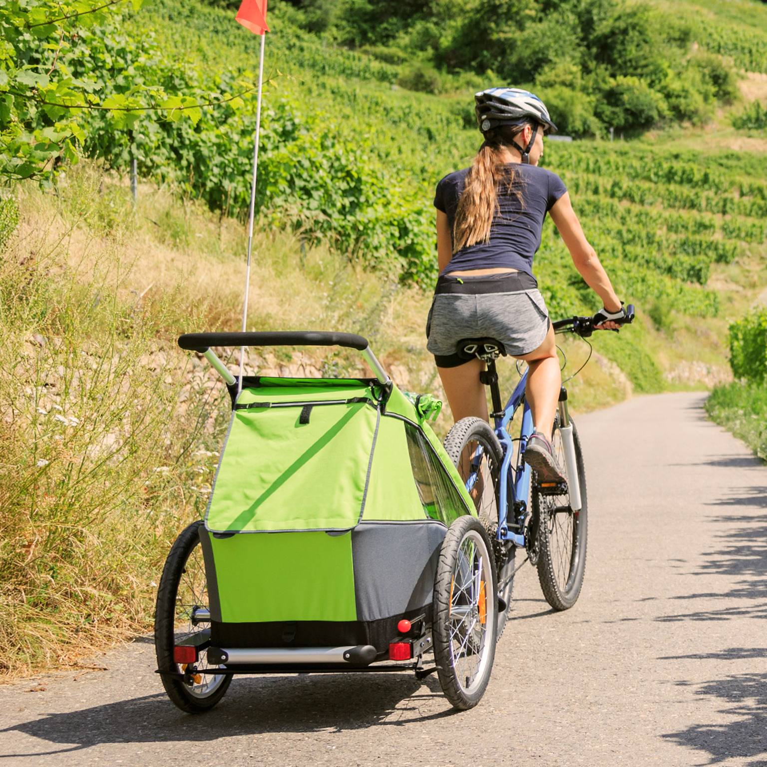 Eine Frau fährt mit dem Fahrrad auf einem Weg durch üppige Weinberge und zieht einen grünen Kinderanhänger hinter sich her. Sie trägt einen Helm und lässige Sportkleidung.