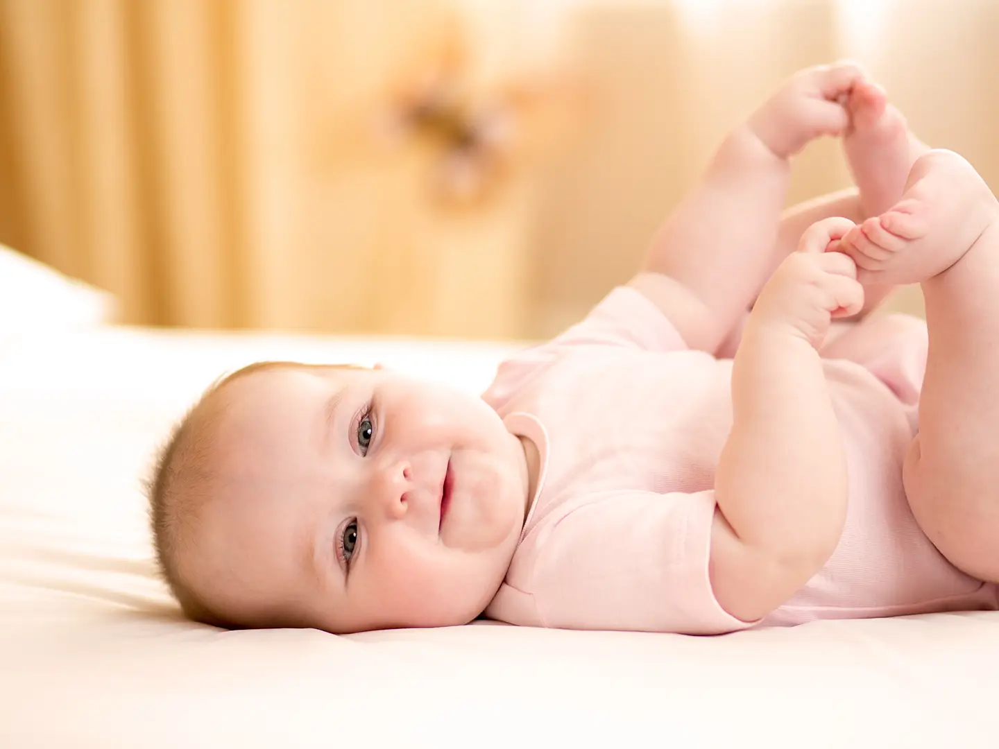 Lächelndes Baby in einem rosa Strampler, das auf einem Bett liegt und seine Füße mit beiden Händen hält, vor einem weichen, unscharfen Hintergrund.