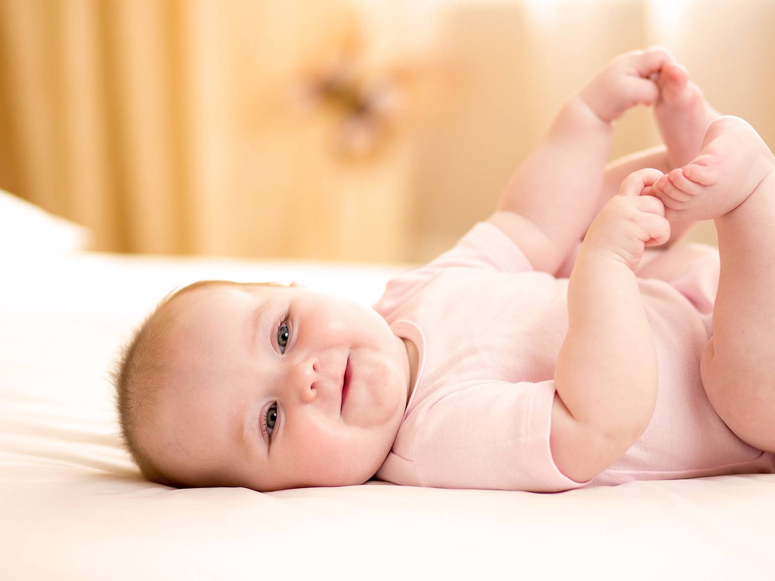 Lächelndes Baby in einem rosa Strampler, das auf einem Bett liegt und seine Füße mit beiden Händen hält, vor einem weichen, unscharfen Hintergrund.