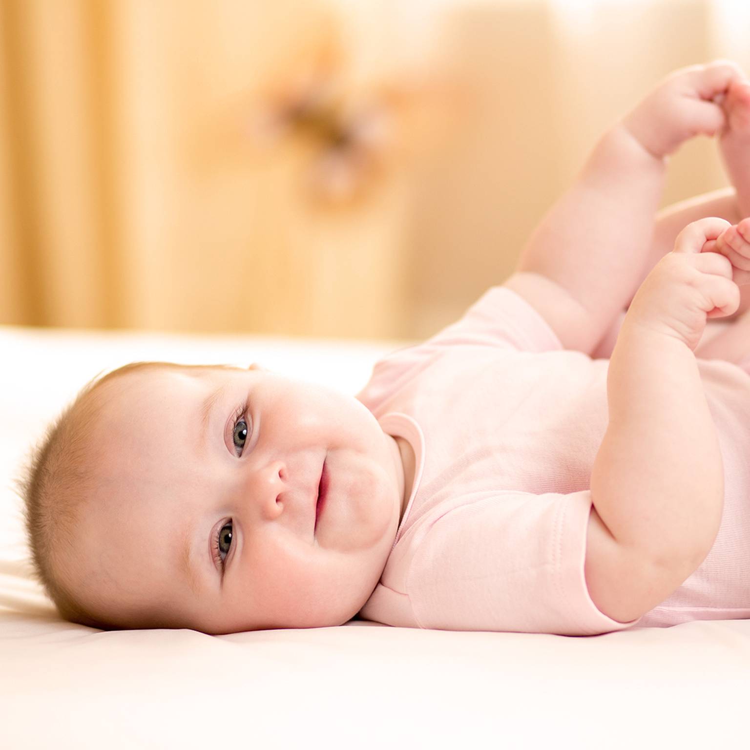 Lächelndes Baby in einem rosa Strampler, das auf einem Bett liegt und seine Füße mit beiden Händen hält, vor einem weichen, unscharfen Hintergrund.
