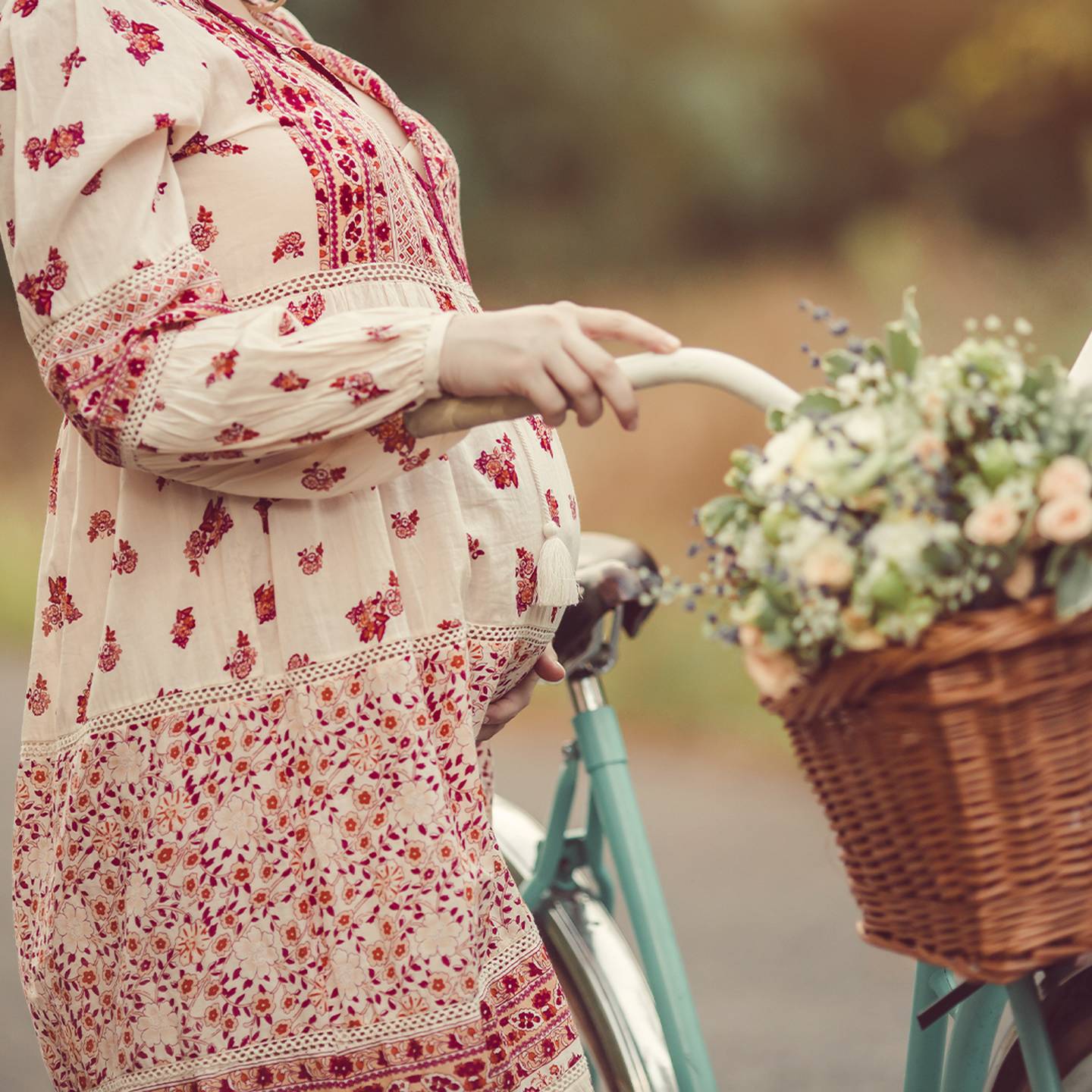 Schwangere Frau im geblümten Kleid steht mit einem Fahrrad, auf dem sich ein Korb voller Blumen und Brot befindet, auf einer Landstraße.