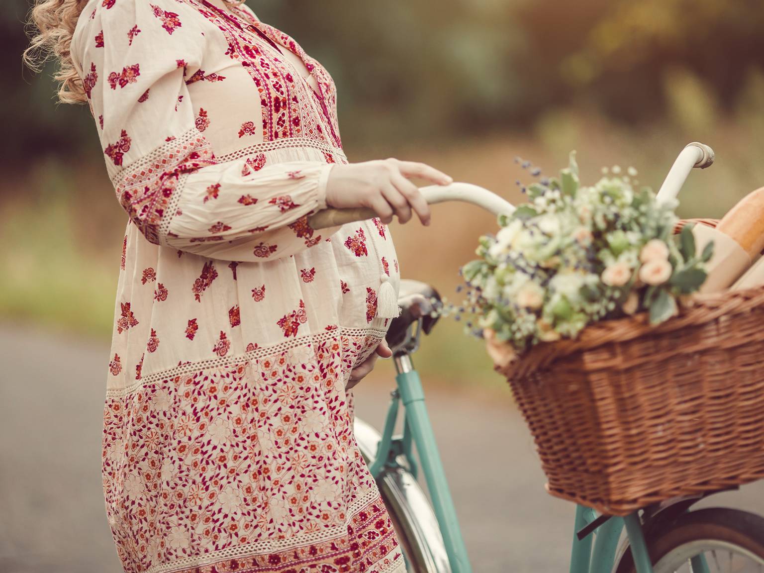 Schwangere Frau im geblümten Kleid steht mit einem Fahrrad, auf dem sich ein Korb voller Blumen und Brot befindet, auf einer Landstraße.