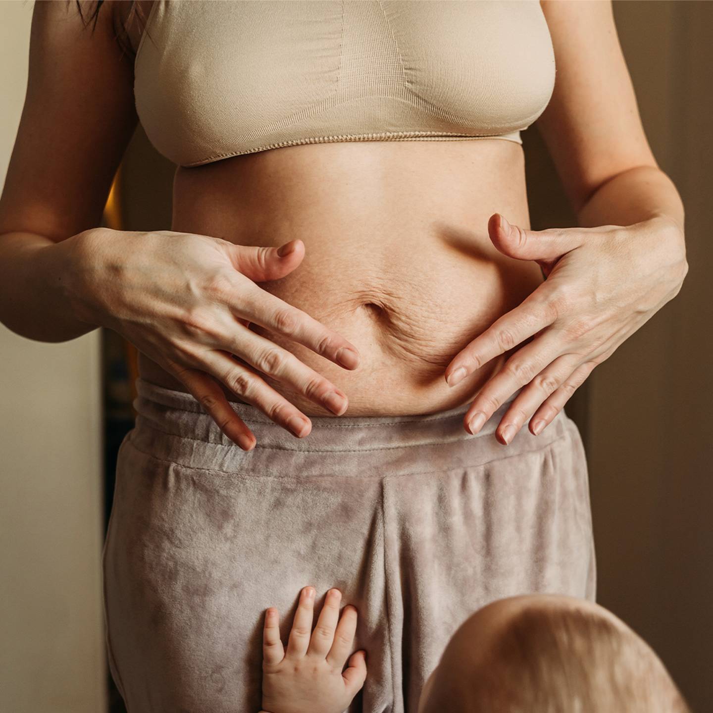 Eine Frau in einem beigen Oberteil zeigt die Haut ihres postpartalen Bauches, während ein Baby ihre Hose berührt. Die Szene vermittelt ein Gefühl von Mutterschaft und Körperfreundlichkeit.