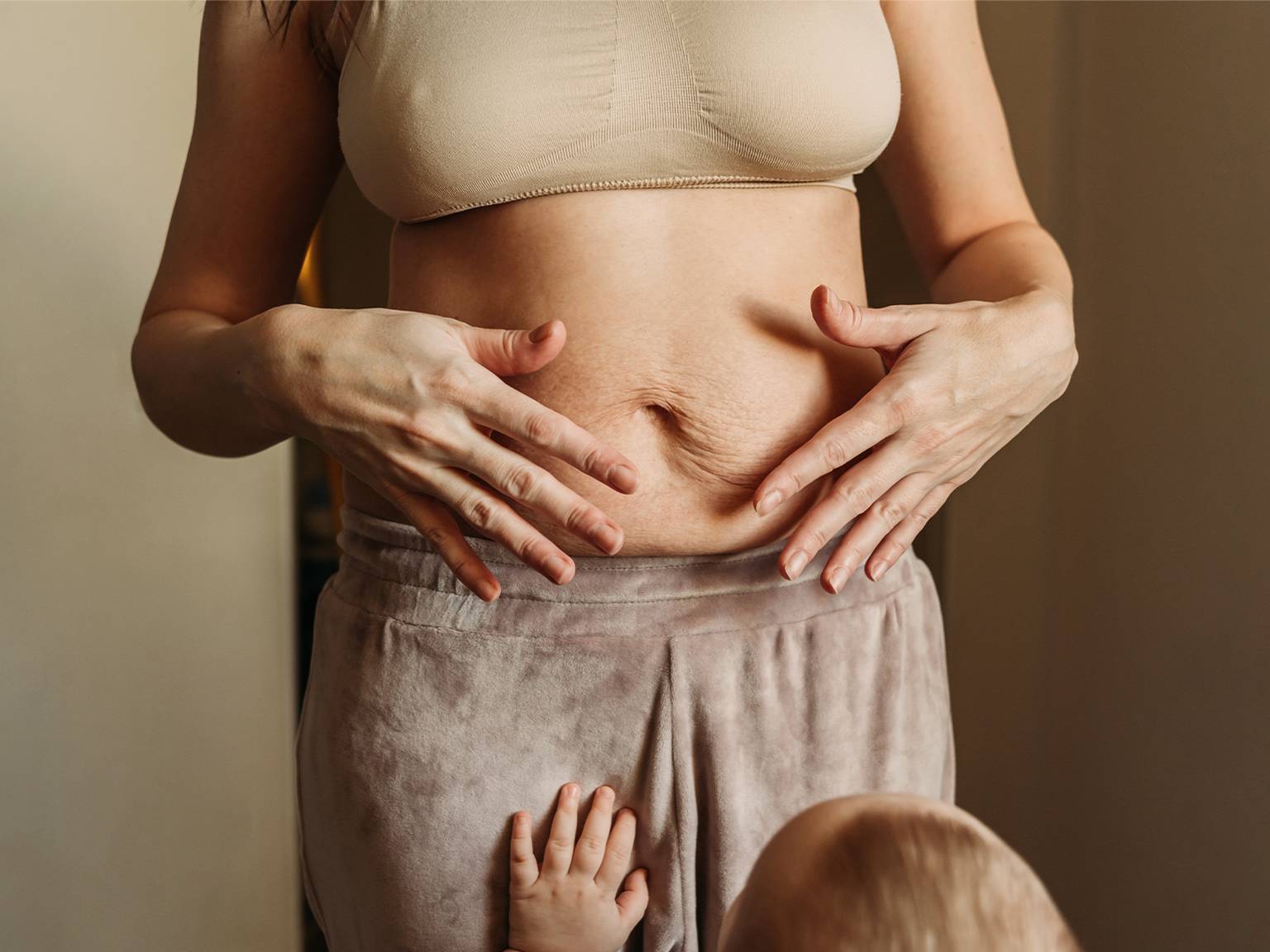 Eine Frau in einem beigen Oberteil zeigt die Haut ihres postpartalen Bauches, während ein Baby ihre Hose berührt. Die Szene vermittelt ein Gefühl von Mutterschaft und Körperfreundlichkeit.