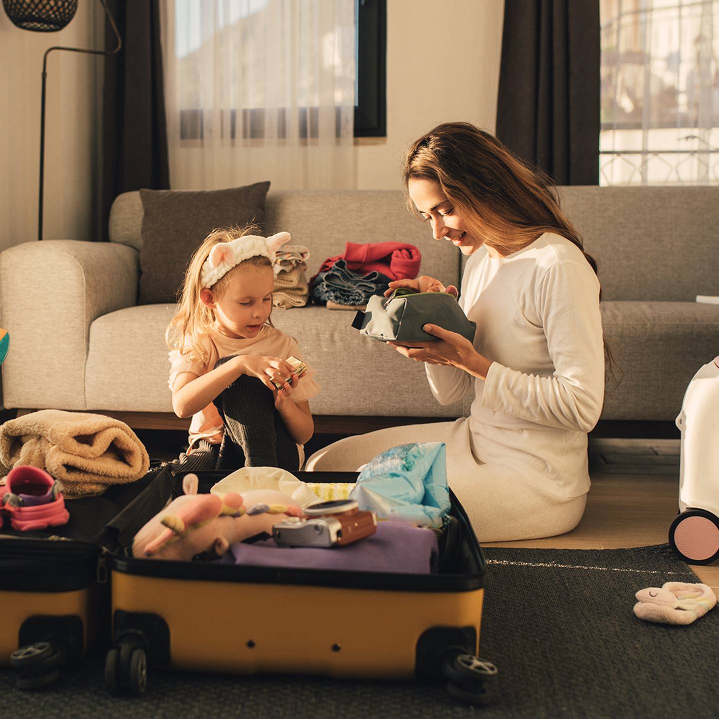 Eine Frau und ein Kind sitzen auf dem Boden und packen einen Koffer, umgeben von Kleidung und Spielsachen in einem gemütlichen Wohnzimmer.