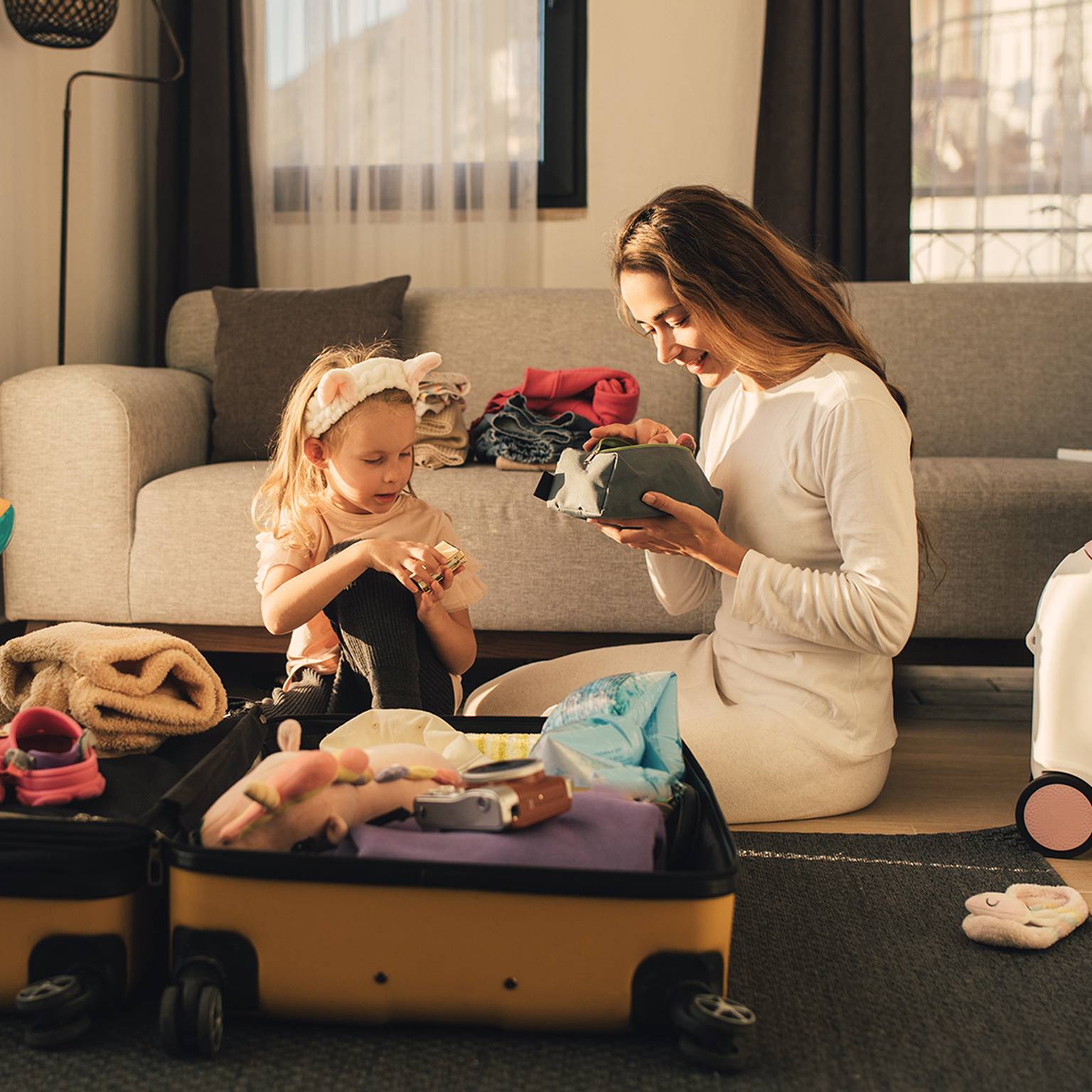 Eine Frau und ein Kind sitzen auf dem Boden und packen einen Koffer, umgeben von Kleidung und Spielsachen in einem gemütlichen Wohnzimmer.