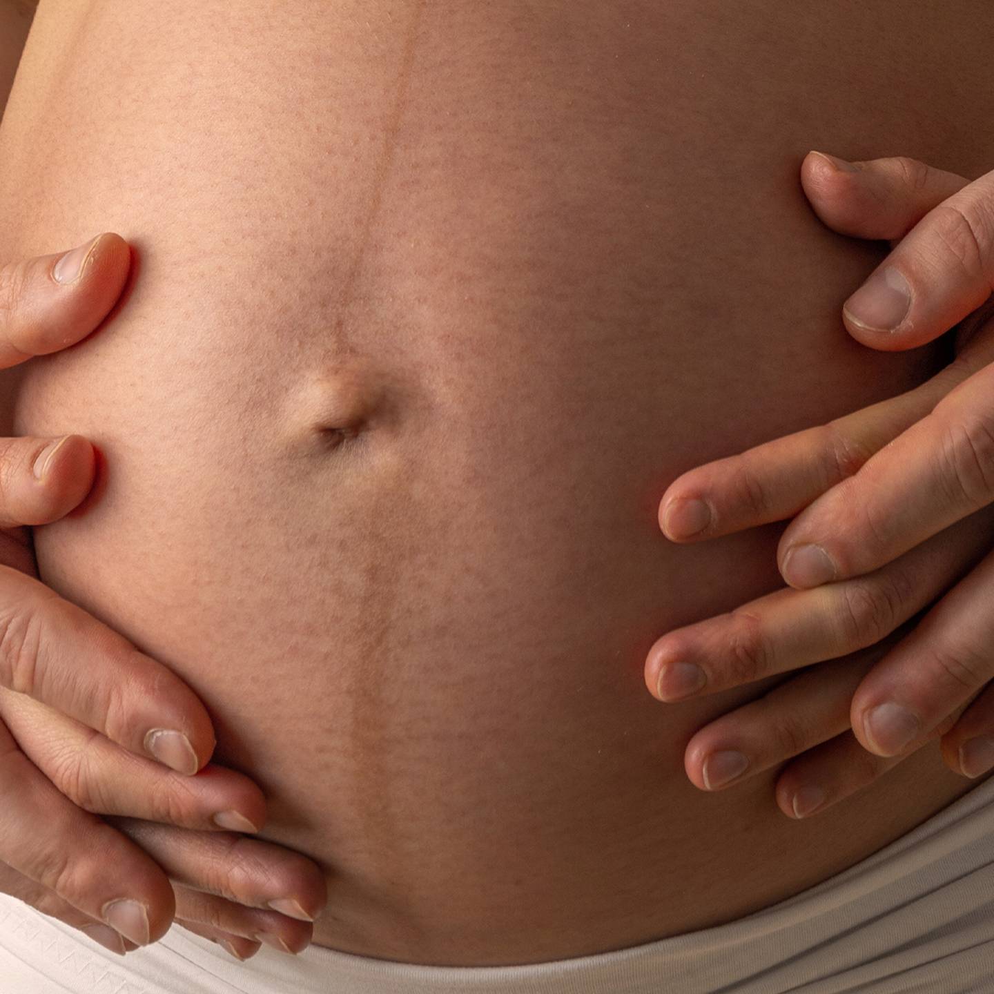 Nahaufnahme von Händen, die sanft auf den Bauch einer Schwangeren gelegt werden. Eine schwache Linea nigra ist zu sehen.