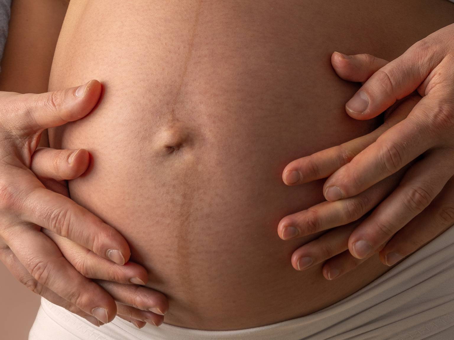 Nahaufnahme von Händen, die sanft auf den Bauch einer Schwangeren gelegt werden. Eine schwache Linea nigra ist zu sehen.