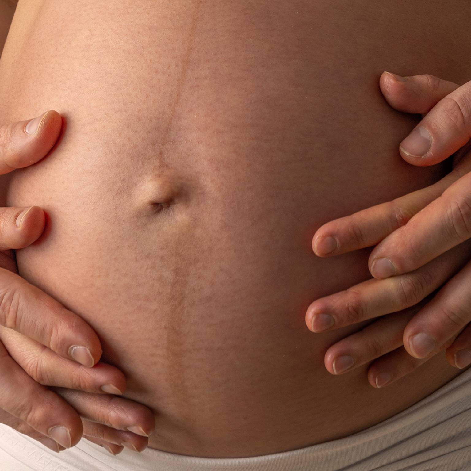 Nahaufnahme von Händen, die sanft auf den Bauch einer Schwangeren gelegt werden. Eine schwache Linea nigra ist zu sehen.