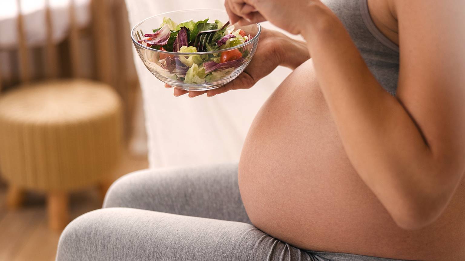 Schwangere Frau in grauer Kleidung sitzt und isst einen frischen Salat aus einer Glasschüssel, im Hintergrund ist ein Kinderbett zu sehen.