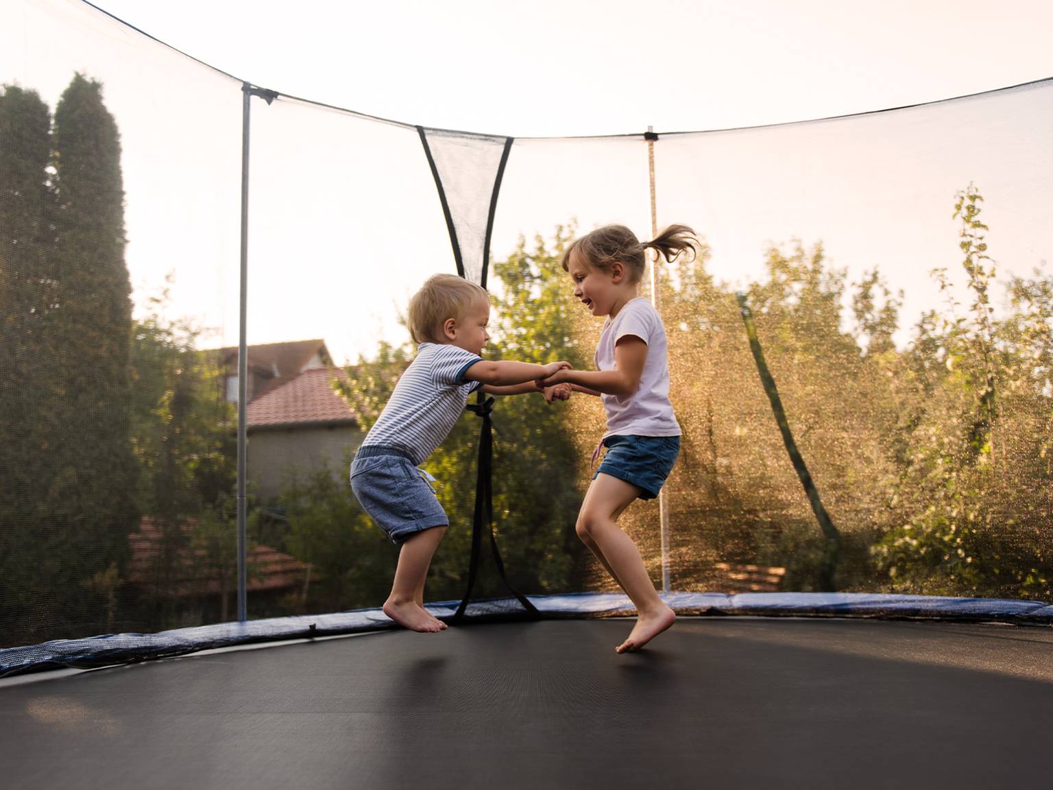 Zwei Kinder hüpfen vergnügt auf einem Trampolin in einem Garten, der von Bäumen umgeben ist. Sie halten sich an den Händen und lächeln sich an.