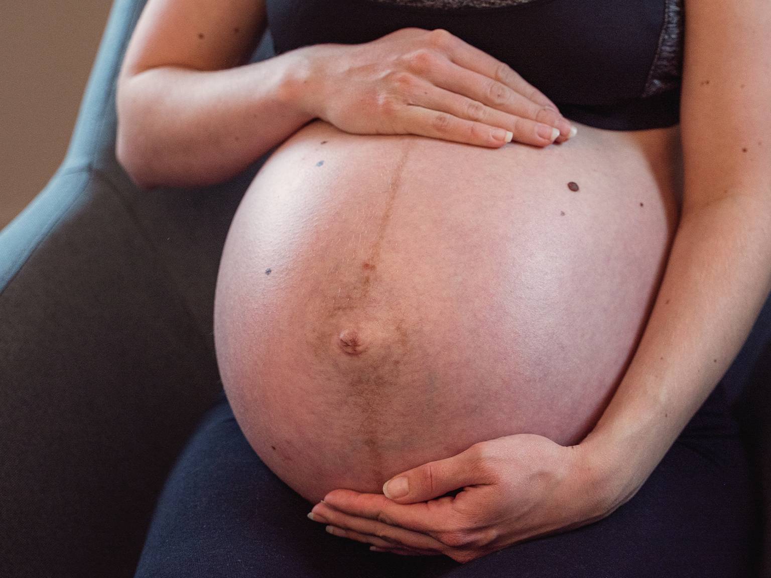 Schwangere sitzt, hält ihren großen Bauch mit beiden Händen und trägt ein schwarzes Outfit.