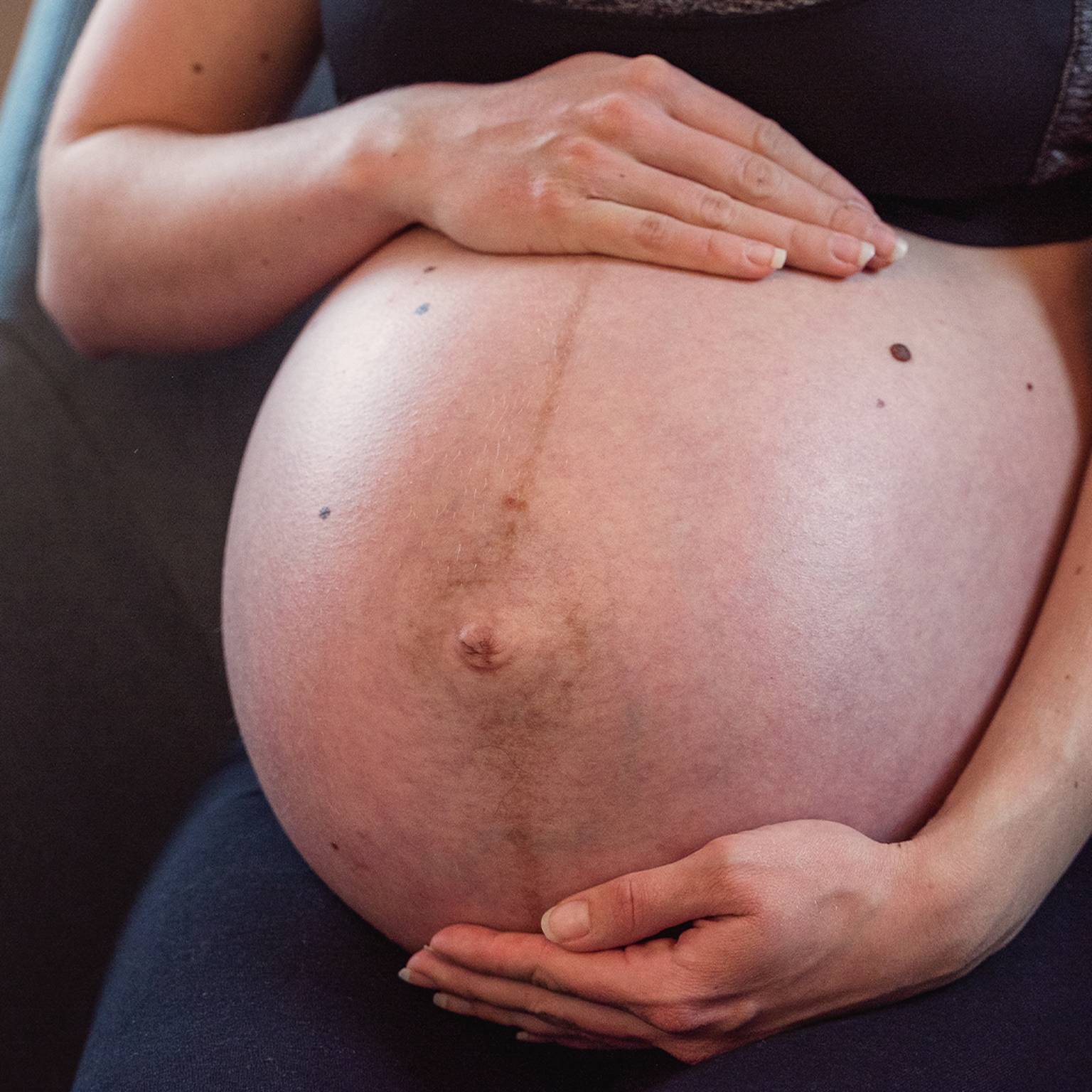 Schwangere sitzt, hält ihren großen Bauch mit beiden Händen und trägt ein schwarzes Outfit.