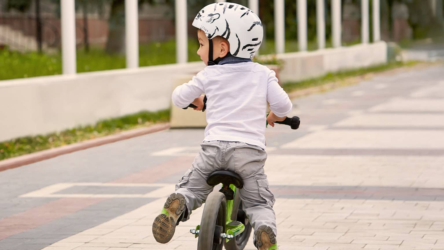 Ein Kind mit Helm fährt mit einem Laufrad auf einem gepflasterten Weg, umgeben von Grün und weißen Säulen.