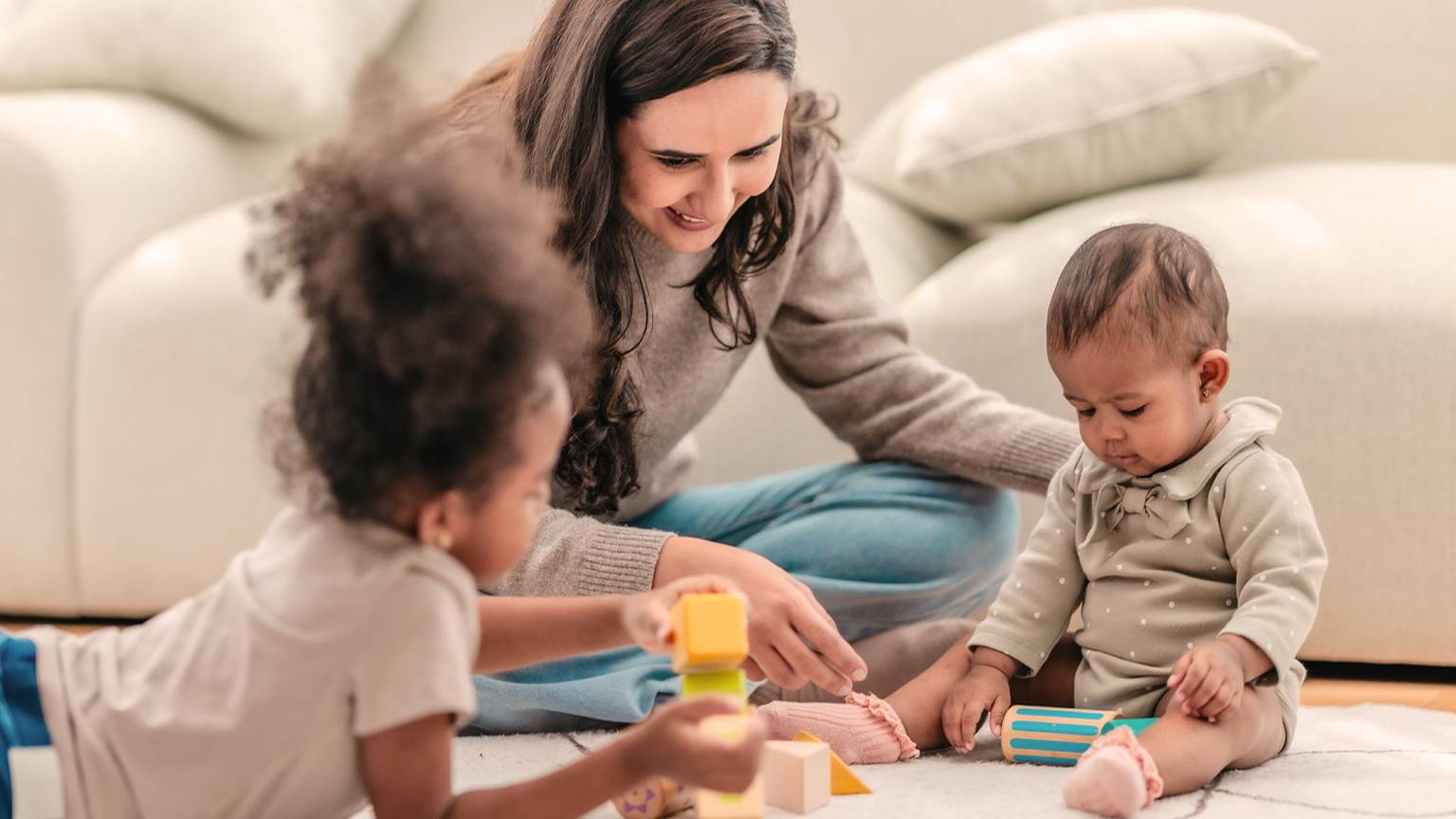 Eine Frau und zwei kleine Kinder spielen in einem gemütlichen Wohnzimmer auf einem Teppichboden mit Bauklötzen.