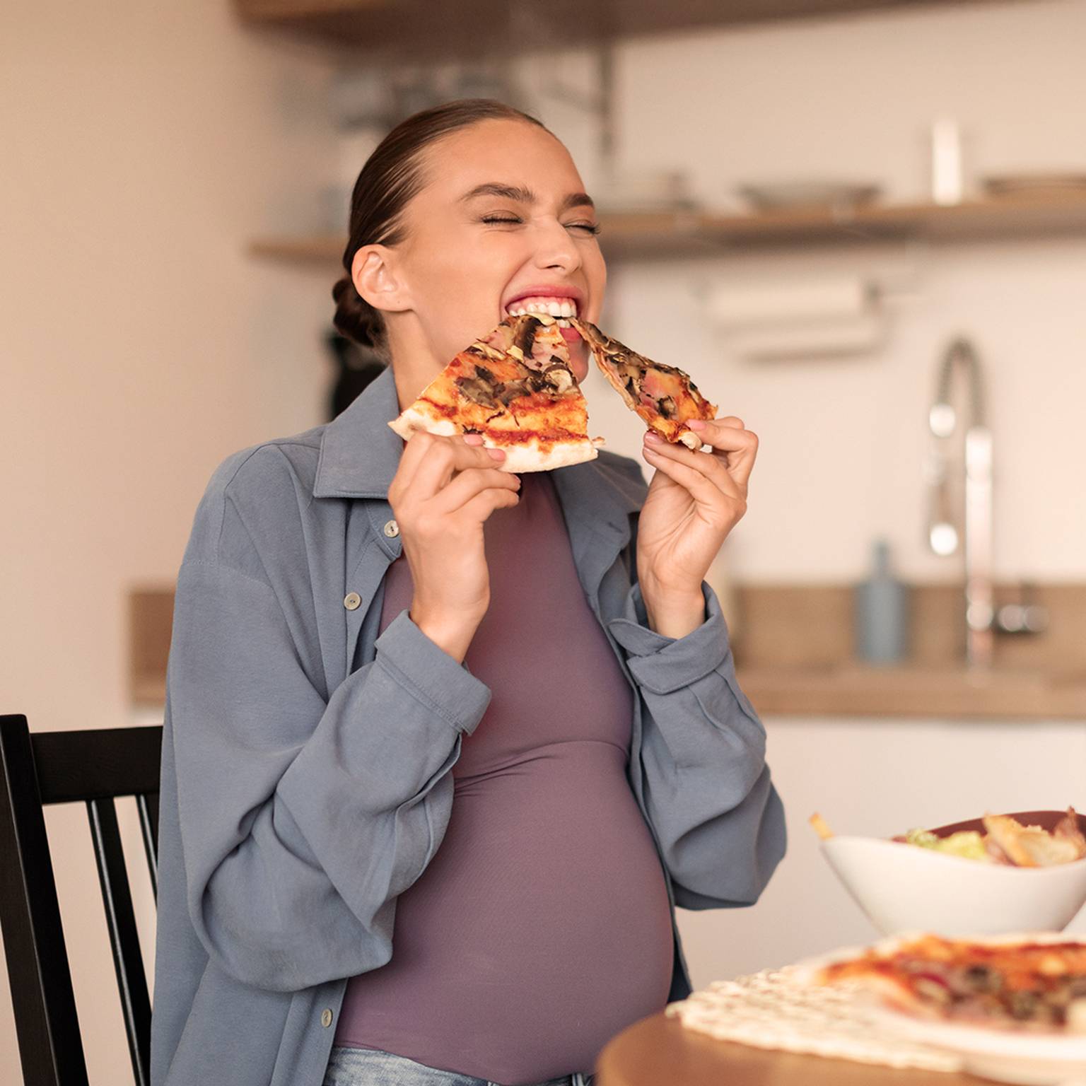Schwangere Frau isst freudig zwei Stücke Pizza an einem Küchentisch, in der Nähe stehen eine Salatschüssel und andere Lebensmittel.