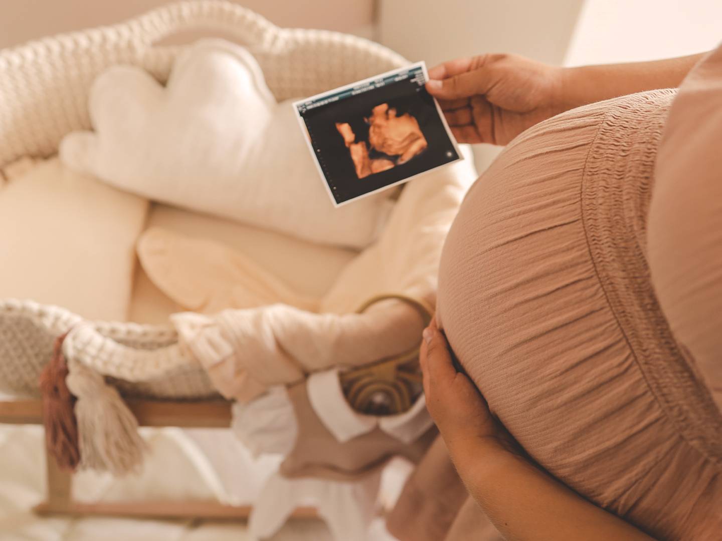 Schwangere Frau hält ein Ultraschallbild in der Hand und steht neben einer Babywiege mit weicher Bettwäsche und einem wolkenförmigen Kissen.