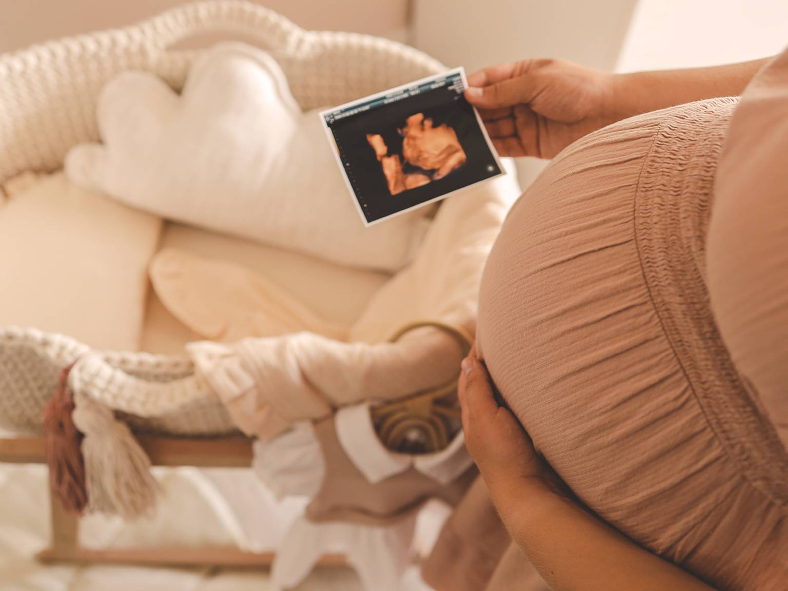 Schwangere Frau hält ein Ultraschallbild in der Hand und steht neben einer Babywiege mit weicher Bettwäsche und einem wolkenförmigen Kissen.