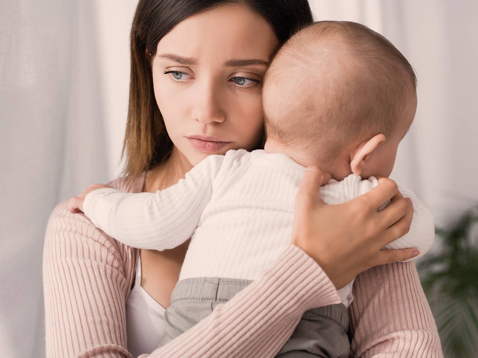 Eine besorgte Frau hält ein Baby im Arm und schaut nachdenklich weg. Das Baby ruht mit dem Kopf auf ihrer Schulter. Beide tragen helle Kleidung.