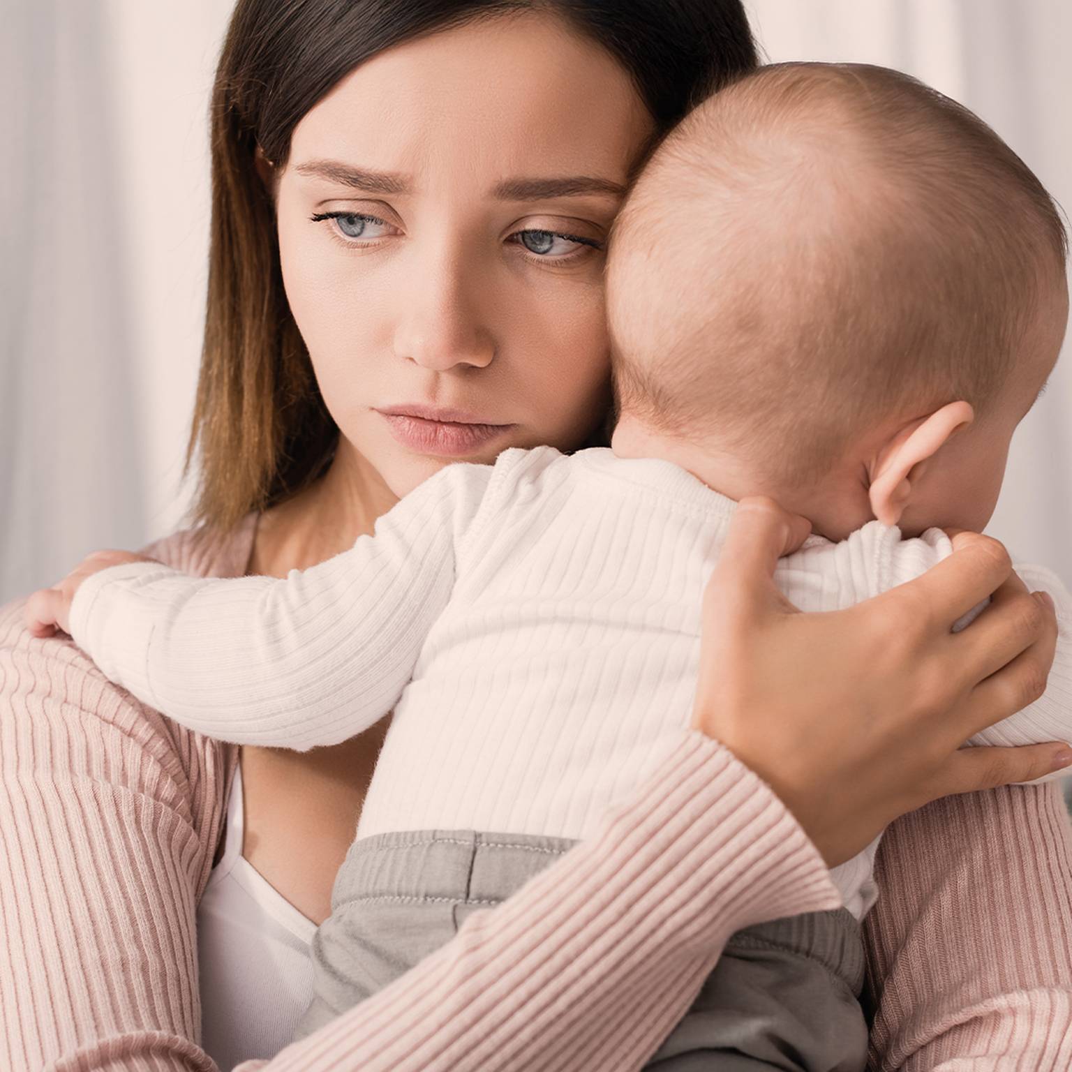 Eine besorgte Frau hält ein Baby im Arm und schaut nachdenklich weg. Das Baby ruht mit dem Kopf auf ihrer Schulter. Beide tragen helle Kleidung.