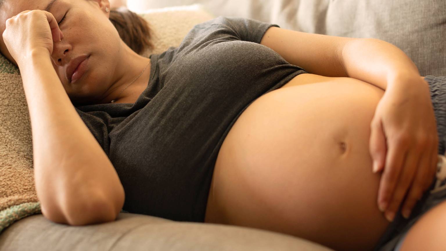 Schwangere Frau ruht auf einer Couch, die Augen geschlossen, die Hand auf der Stirn, trägt ein graues Hemd und Shorts und zeigt einen entspannten und müden Gesichtsausdruck.