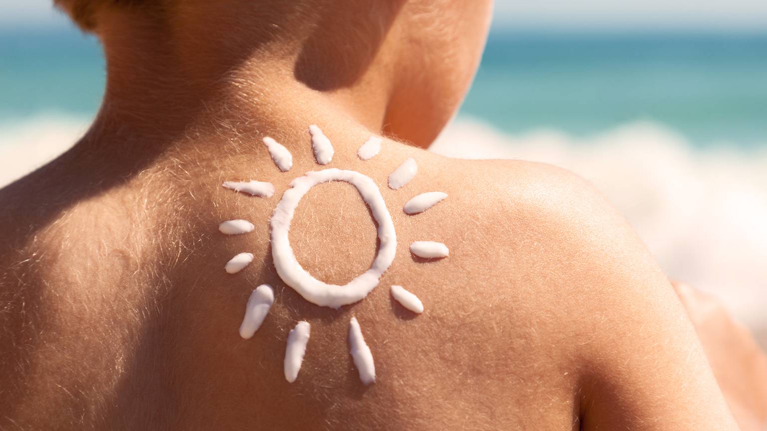 Rücken eines Kindes mit gezeichneter Sonne in Sonnencreme-Lotion, Strand und Meer im Hintergrund.