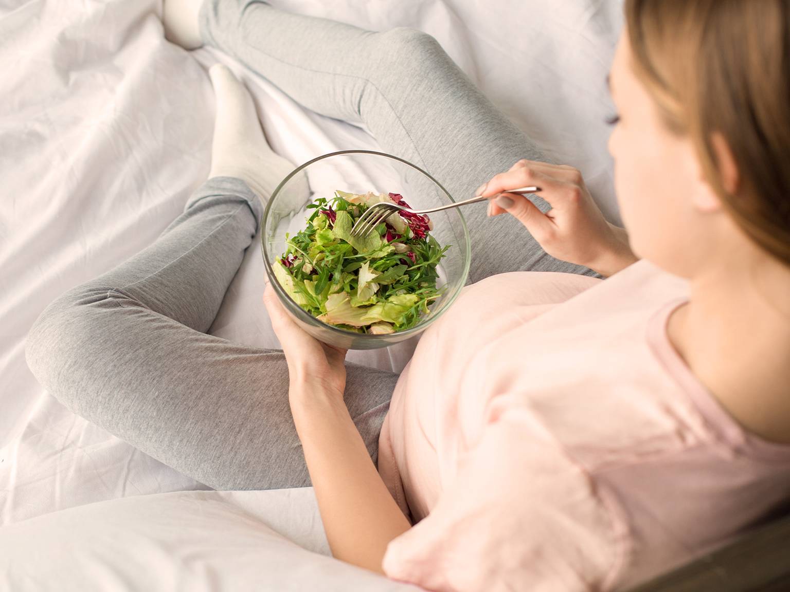 Person in Freizeitkleidung sitzt auf einem Bett, hält eine Schüssel mit frischem Salat und isst mit einer Gabel.