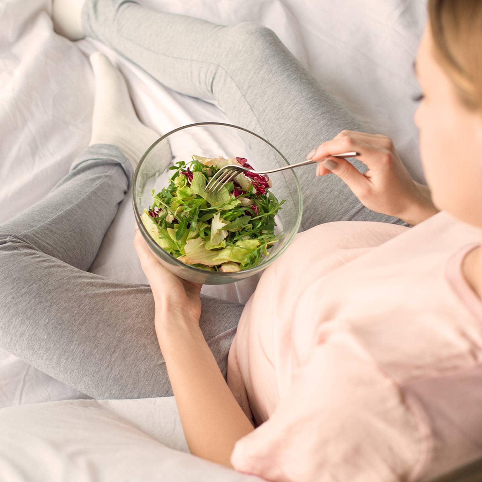 Person in Freizeitkleidung sitzt auf einem Bett, hält eine Schüssel mit frischem Salat und isst mit einer Gabel.