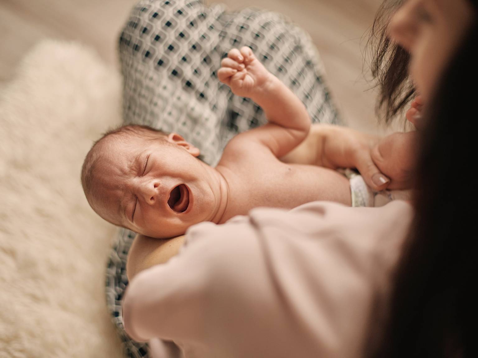 Ein weinendes Baby liegt in den Armen eines Erwachsenen, der eine Windel trägt. Der Erwachsene ist teilweise sichtbar und hält das Baby sanft.